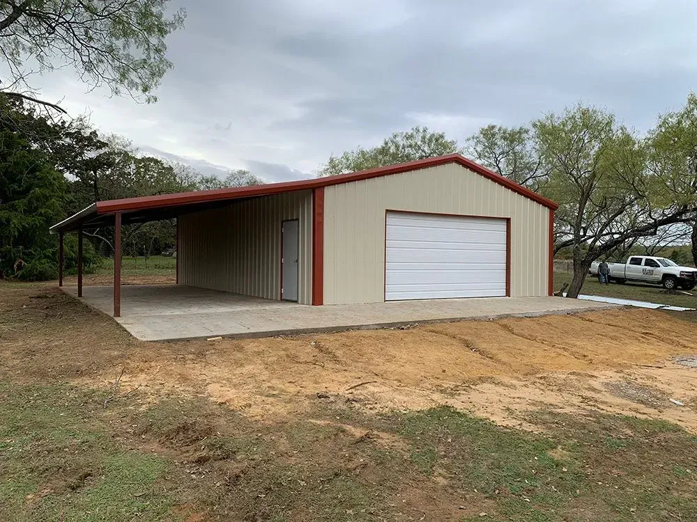 A metal-sided workshop with a covered side patio, a white garage door, and a red roof, situated on a grassy lot.
