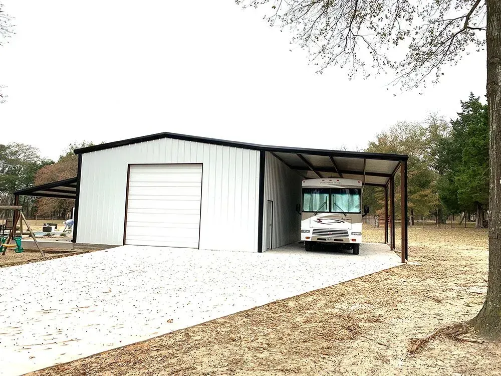 A white metal workshop with a large roll-up door and an open carport housing a motorhome on a gravel lot.