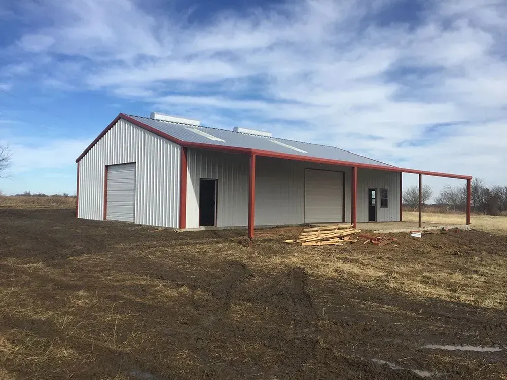 A modern metal workshop building with a grey roof and side, red trim, and covered patio under a blue, cloudy sky.