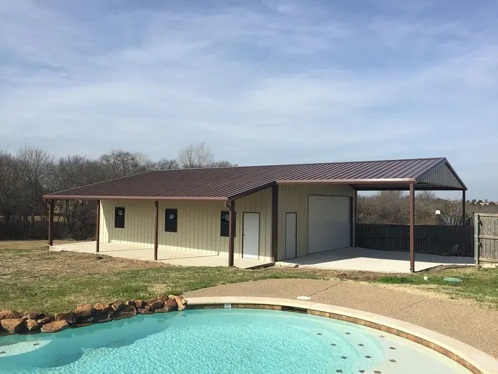 A metal building with a garage and attached carport sits on a grassy lot next to a blue swimming pool under a blue sky.