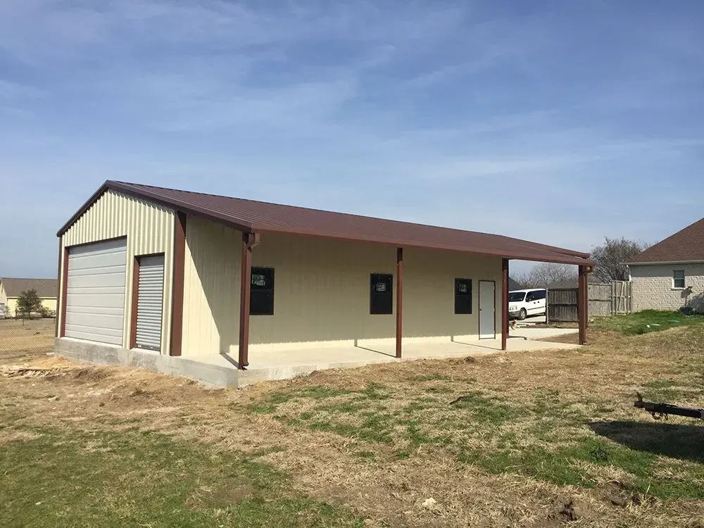 A tan metal shop building with a brown roof, a garage door, side windows, and an open covered porch in a grassy yard.