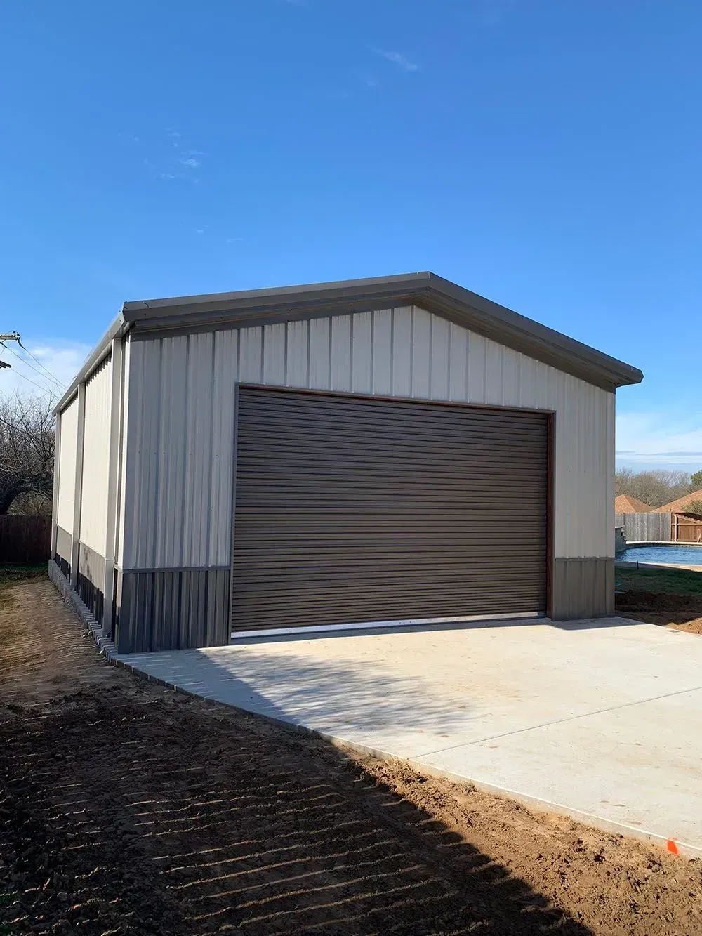 A metal garage with light-colored siding, a dark trim base, and a dark brown roll-up door on a concrete driveway.