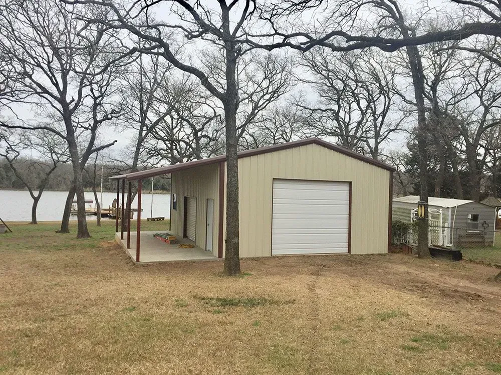 A beige metal garage with a side porch and a white roll-up door, situated on a grassy lot near a lake and trees.