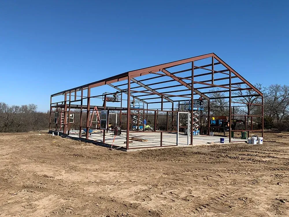 A brown steel frame structure for a building under construction on a dirt lot under a clear blue sky.