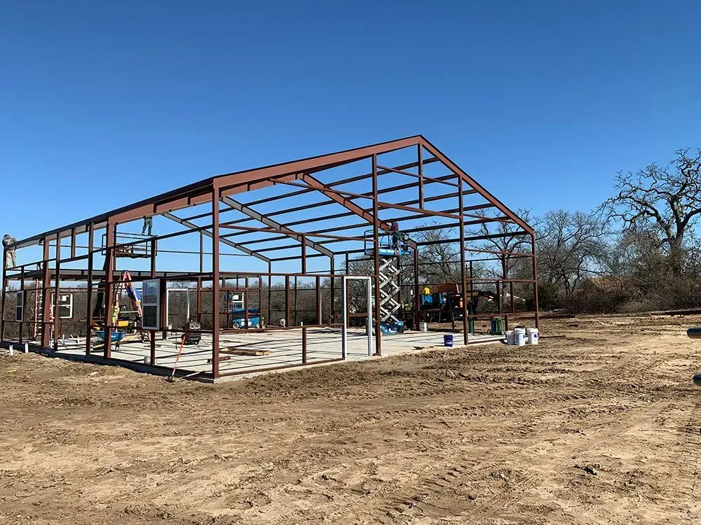 Steel frame of a building under construction on a dirt lot under a clear blue sky.