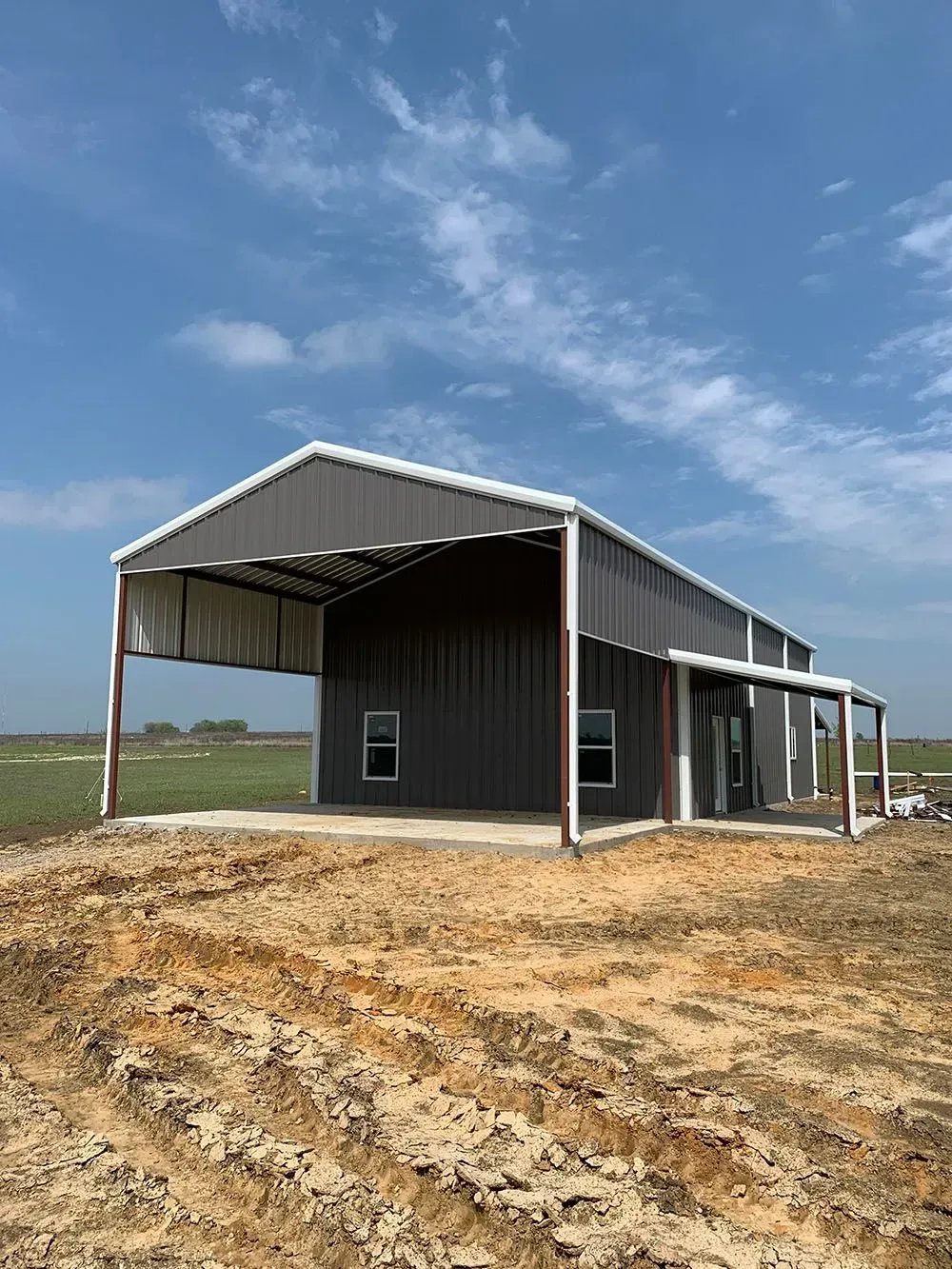 A dark gray metal barn with an open-sided carport under a blue sky, set on a dirt construction lot.