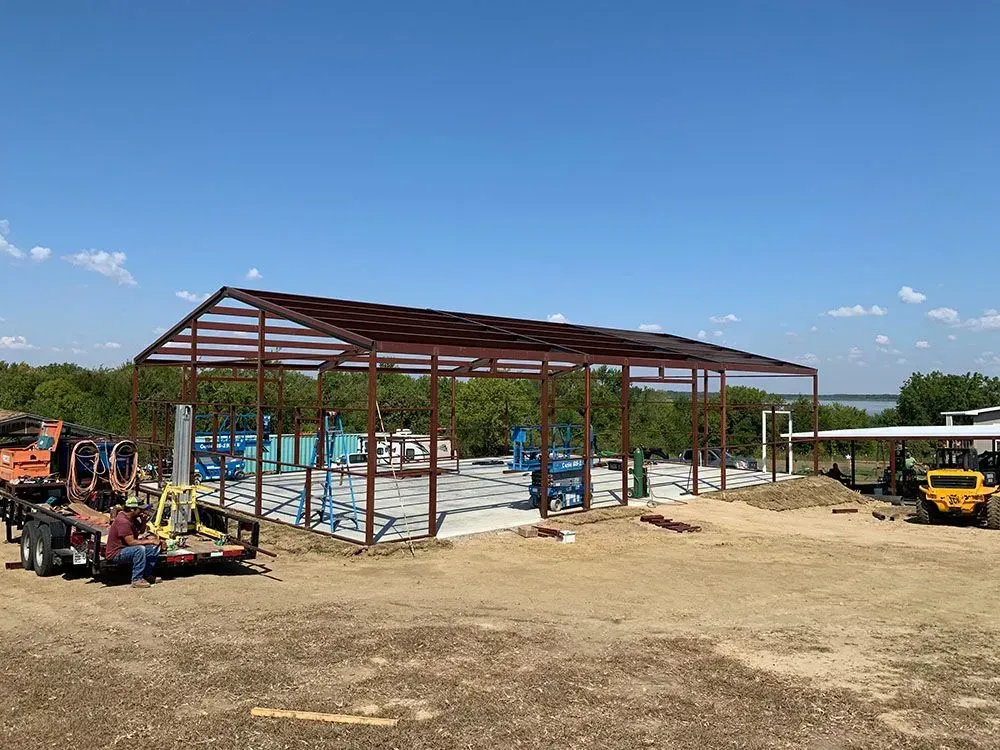 The steel frame of a rectangular metal building stands on a concrete slab in a grassy field under a clear blue sky.