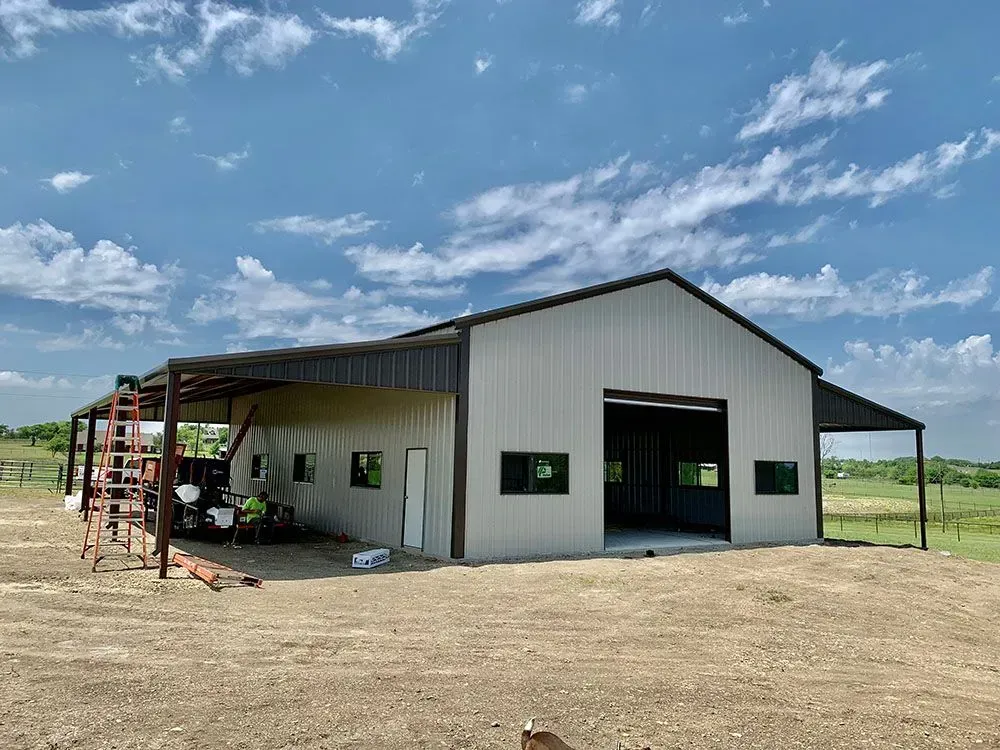 A large, light-colored metal pole barn with dark trim and an open side porch stands on a dirt lot under a blue sky.