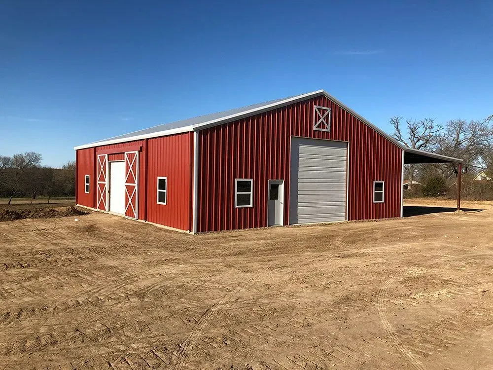 A red metal barn with white trim and a roll-up door, situated on a dirt lot under a clear blue sky.