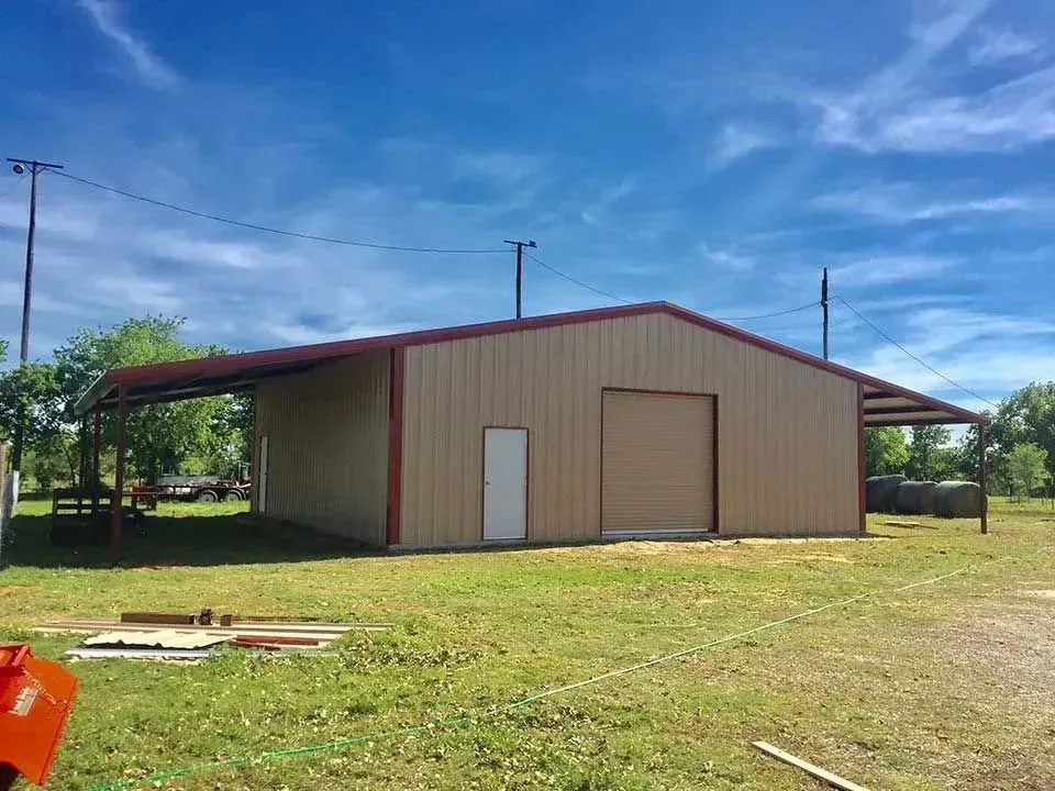 A tan metal building with a red frame, a large roll-up door, a side door, and side roof overhangs in a grassy field.