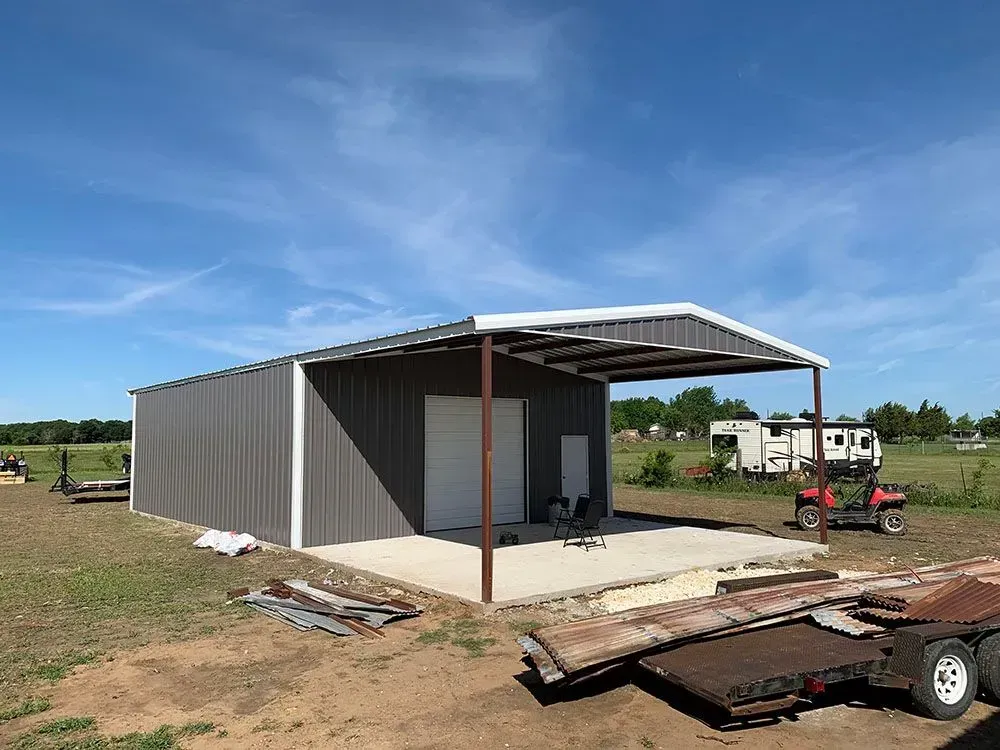 A grey metal barn with an open front patio, set in a sunny, rural outdoor field with a trailer parked nearby.