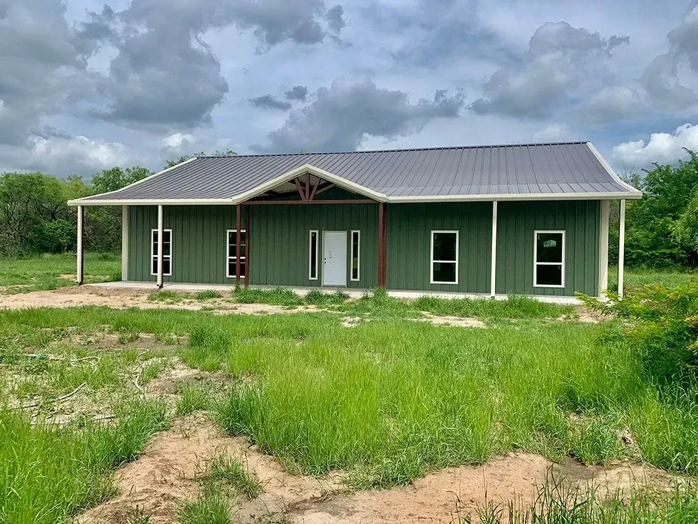 A single-story, green metal-sided house with a gray metal roof, white trim, and a small front porch under a cloudy sky.