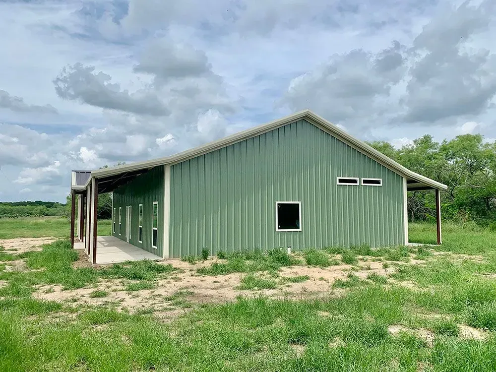 A light green metal building with a gabled roof and covered side porch, set in a grassy field under a cloudy sky.