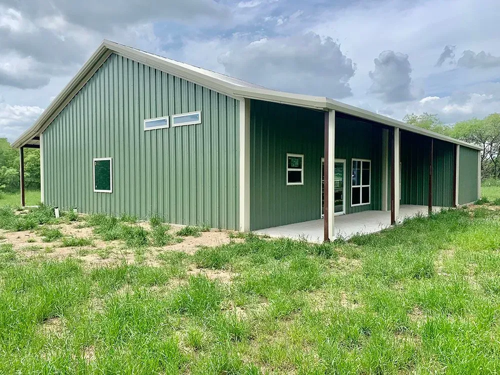 A green metal building with a slanted roof, porch, and windows, set in a grassy field under a cloudy sky.