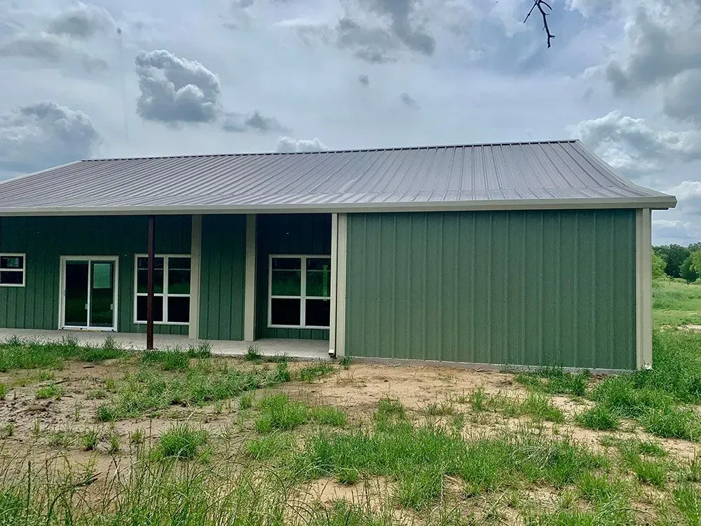 A green, metal-sided barn-style building with a grey roof and glass sliding doors, set in a grassy rural landscape.