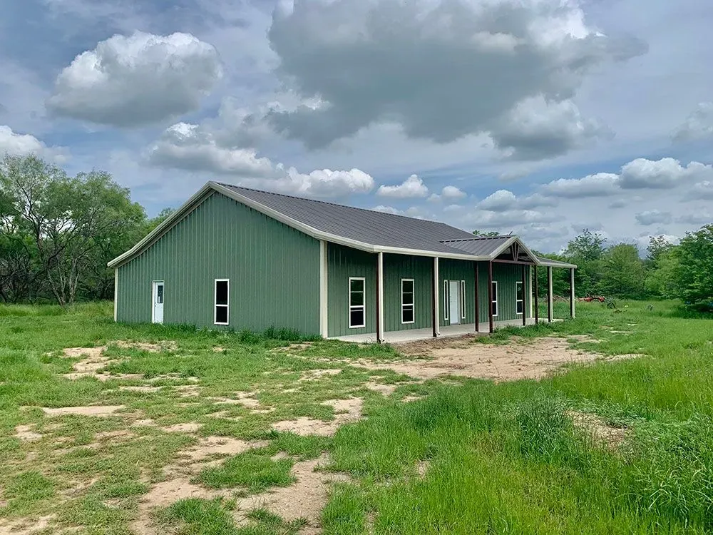 A green metal building with a dark roof and covered porch, set in an open grassy field under a cloudy sky.