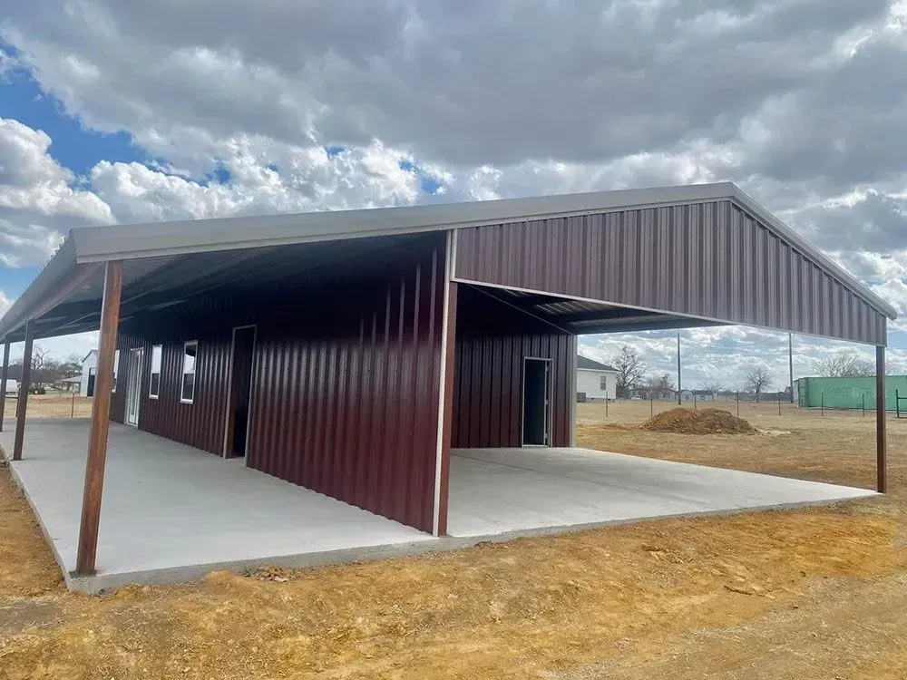 A modern metal barn with a maroon-paneled exterior and a large covered concrete porch under a cloudy sky.