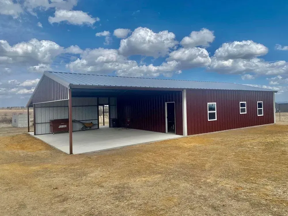 A long, red metal building with a concrete porch and open bay, set in a dry, grassy field under a blue sky with clouds.