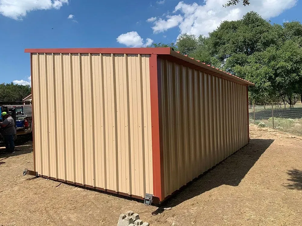 A tan metal storage shed with red trim stands on a dirt lot under a blue sky, with a truck visible in the distance.