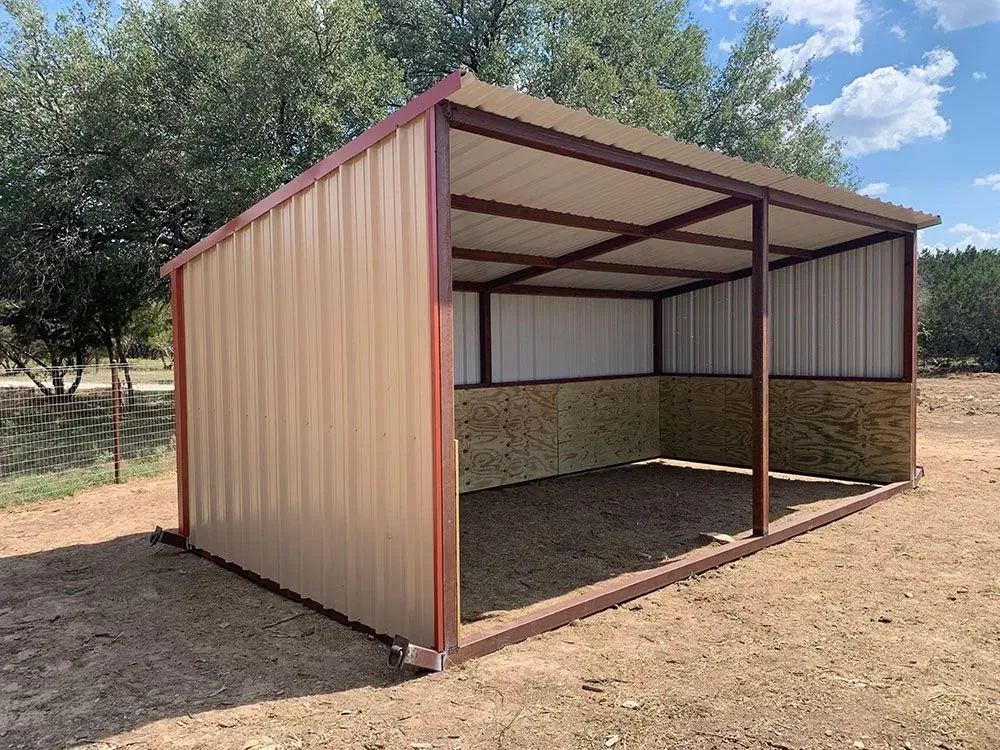 A tan, open-sided metal horse shelter with a sloped roof and wooden interior walls, situated in a dry, grassy outdoor area.