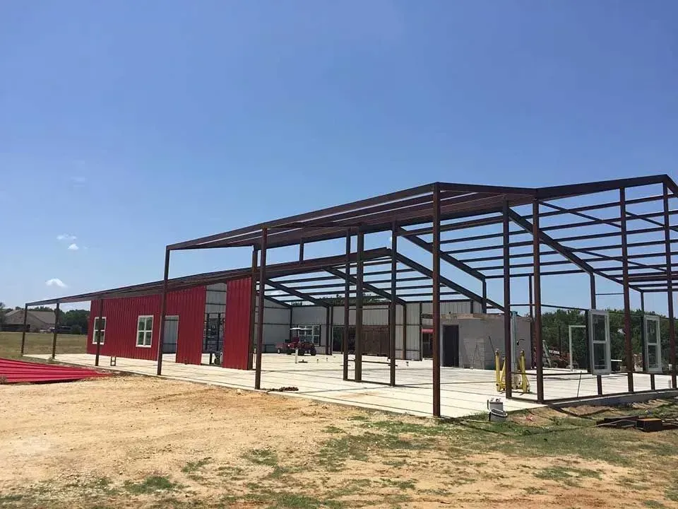 A large, open-air metal building frame under construction with red siding partially installed on a sunny day.