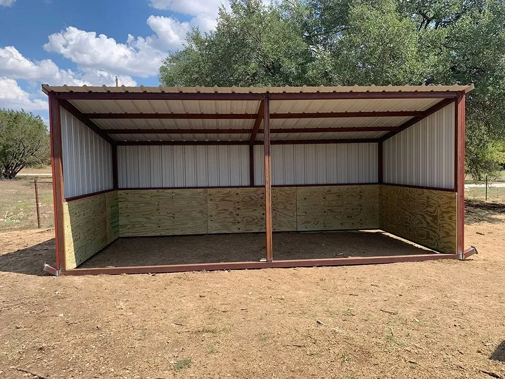 A metal two-stall run-in shelter with corrugated white walls, wood-paneled lower sides, and a brown frame in a grassy field.