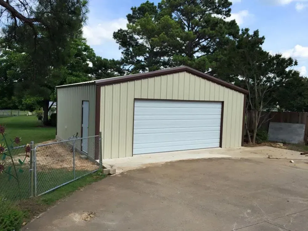 A tan metal detached garage with a white roll-up door and a side entry door, situated on a concrete slab in a yard.