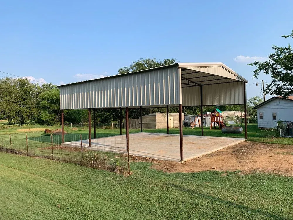 A metal carport with a gabled roof stands on a concrete pad in a grassy yard under a clear blue sky.