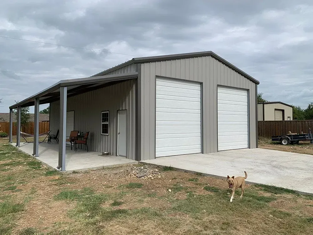 A light gray metal garage with two roll-up doors, a side patio, and a dog walking on the concrete in the foreground.