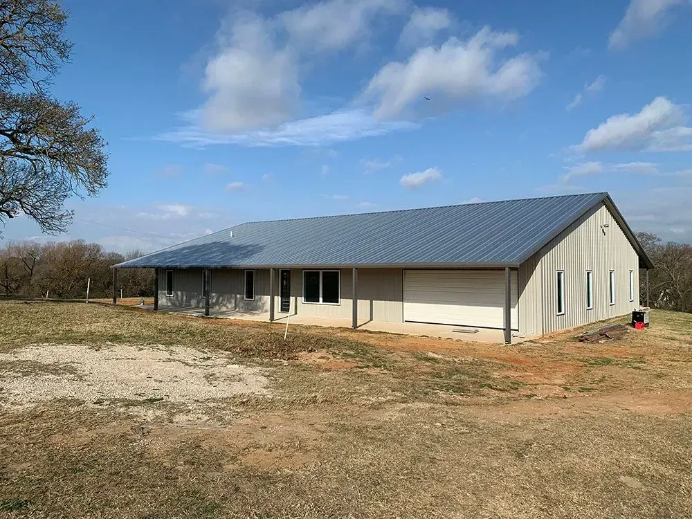 A single-story metal-sided house with a grey roof stands on a dry, grassy rural lot under a blue sky with scattered clouds.