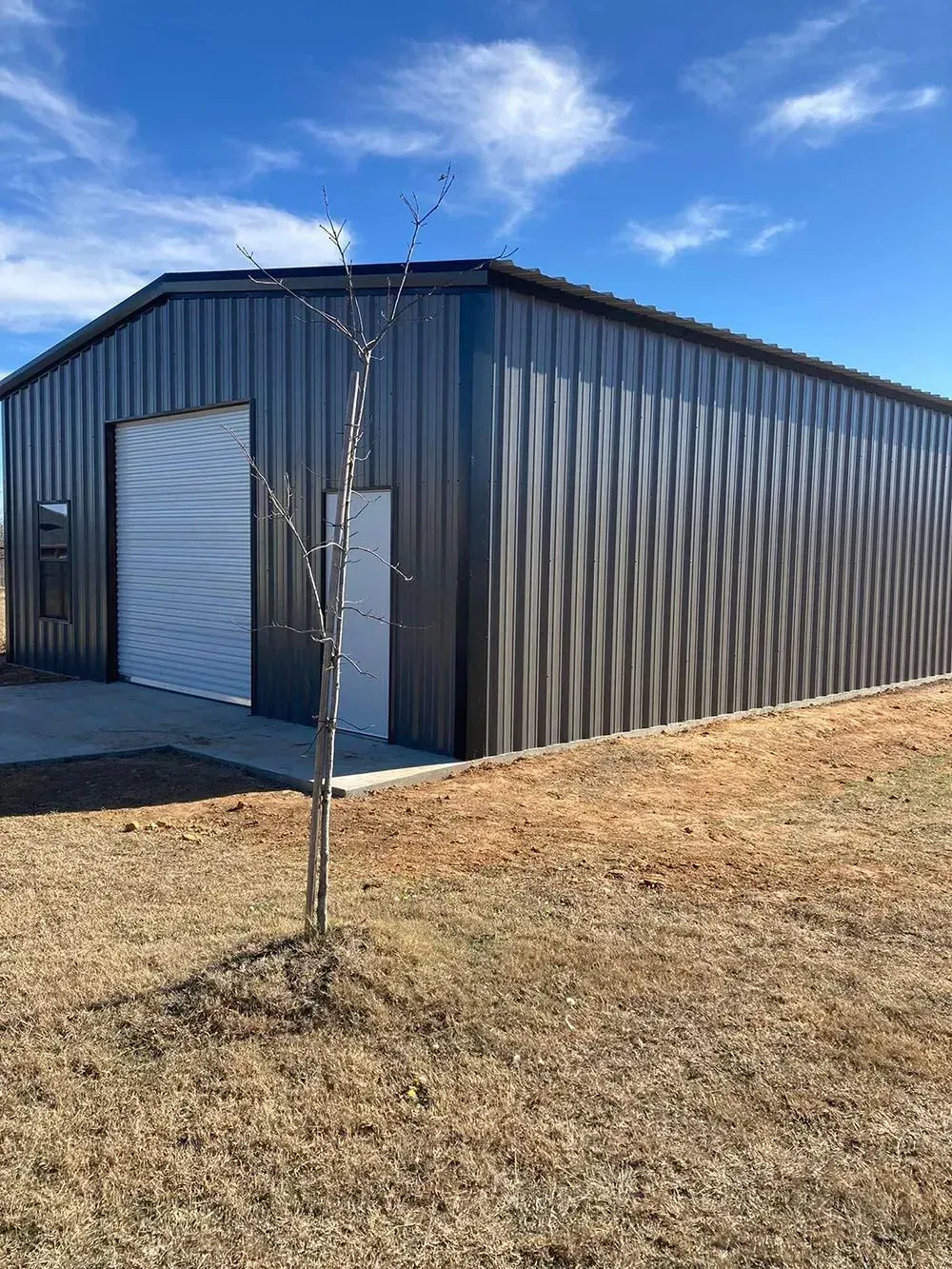 A modern, dark grey metal workshop building with a roll-up door and side entrance, set in a sunny, grassy rural field.