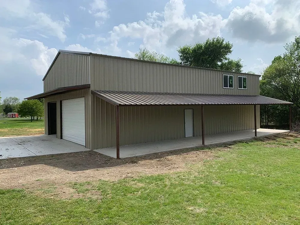 A tan, metal-sided barn with a brown roof, a white garage door, a side entry door, and a full-length covered porch.