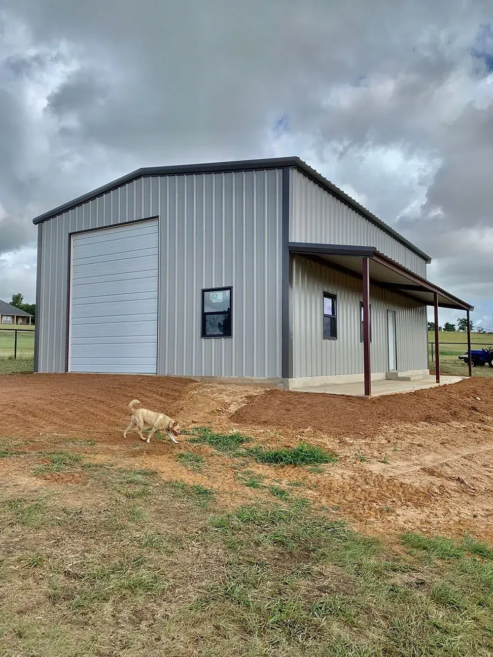 A grey metal barn with a large roll-up door and side porch, located on a grassy hill with a small dog walking in front.