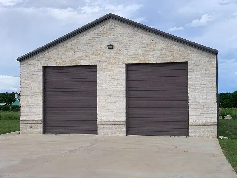 A detached two-car garage with light-colored stone siding and two brown, horizontal-paneled roll-up doors.
