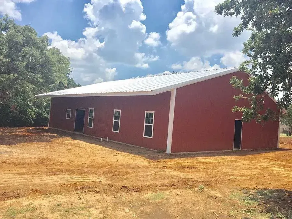 A rectangular, red metal building with a white roof and trim, surrounded by cleared dirt, set against a cloudy blue sky.