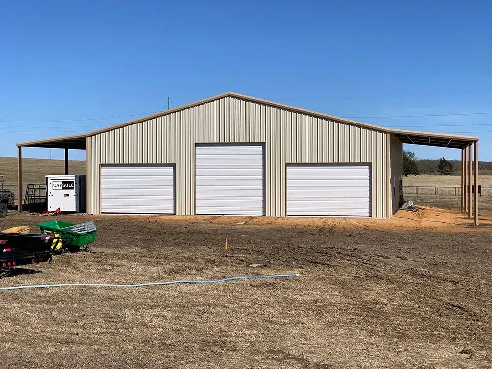 A beige metal-sided barn with three white garage doors and open lean-tos on both sides, set in a rural field.