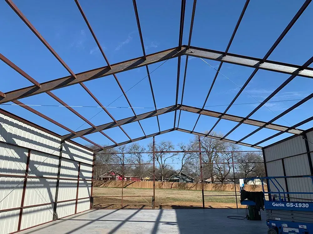 An unfinished metal building frame with a concrete floor, viewed from inside looking out toward a treed field.