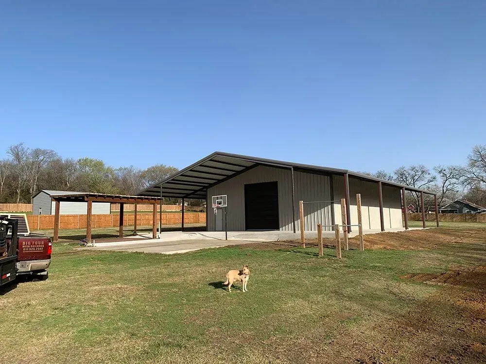 A large light-colored metal barn with a wide front porch and a basketball hoop, set in a grassy yard with a dog nearby.