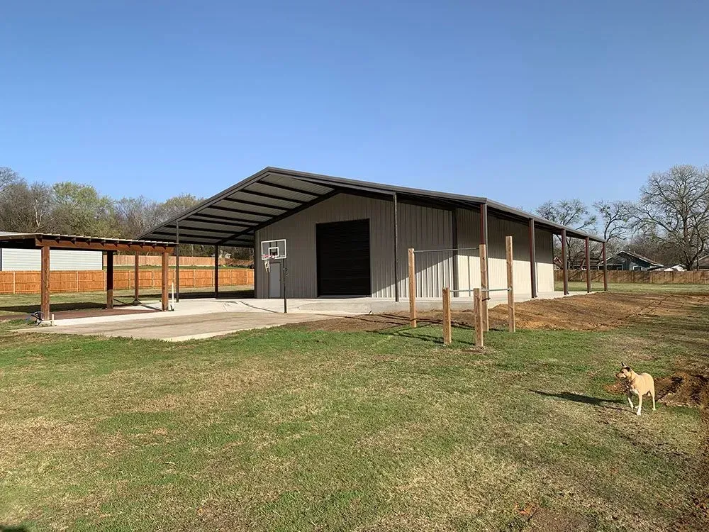 A metal-sided workshop with an overhanging roof, a basketball hoop, and a small dog on a grassy lot.