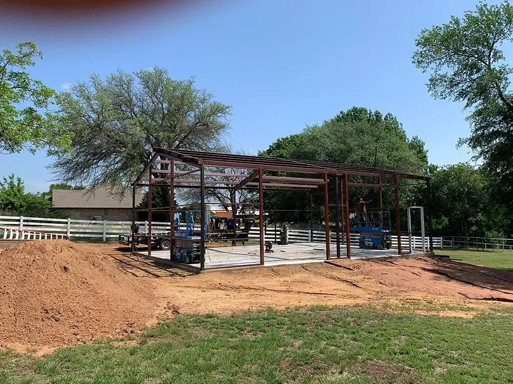 A metal building frame stands on a concrete foundation in a grassy yard, with a large pile of dirt in the foreground.