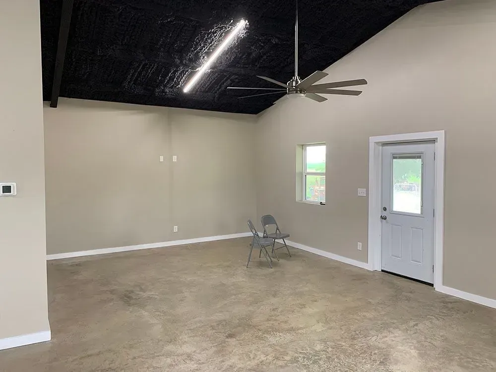 A bright room with beige walls, a black ceiling, a ceiling fan, a window, a white door, and two chairs on concrete floors.