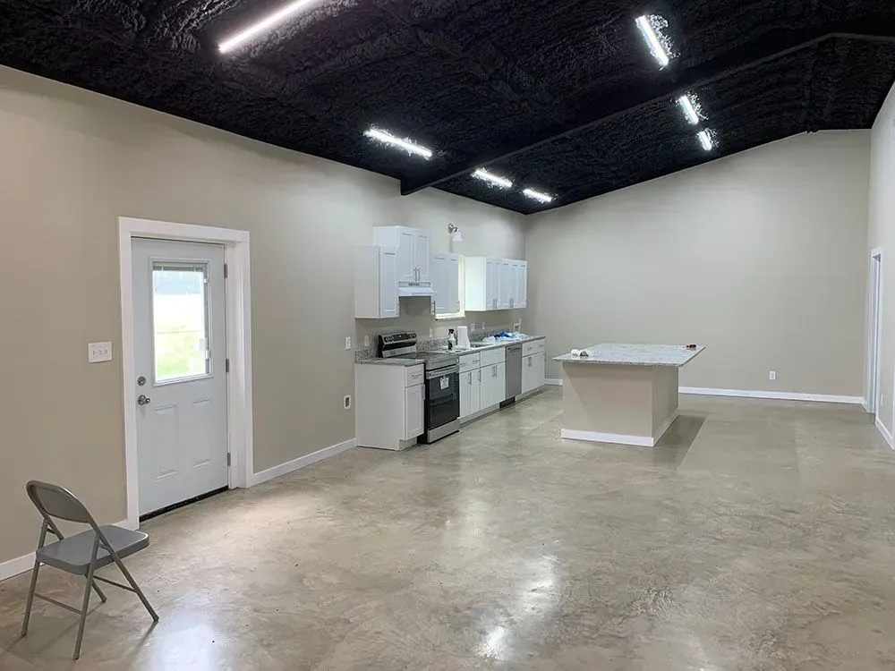 An open-concept room with light walls, a dark ceiling, concrete floors, a small kitchenette, an island, and a folding chair.