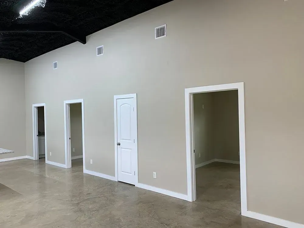 A wide, beige interior wall featuring a black ceiling, concrete floors, three doorways, and four high, small wall vents.