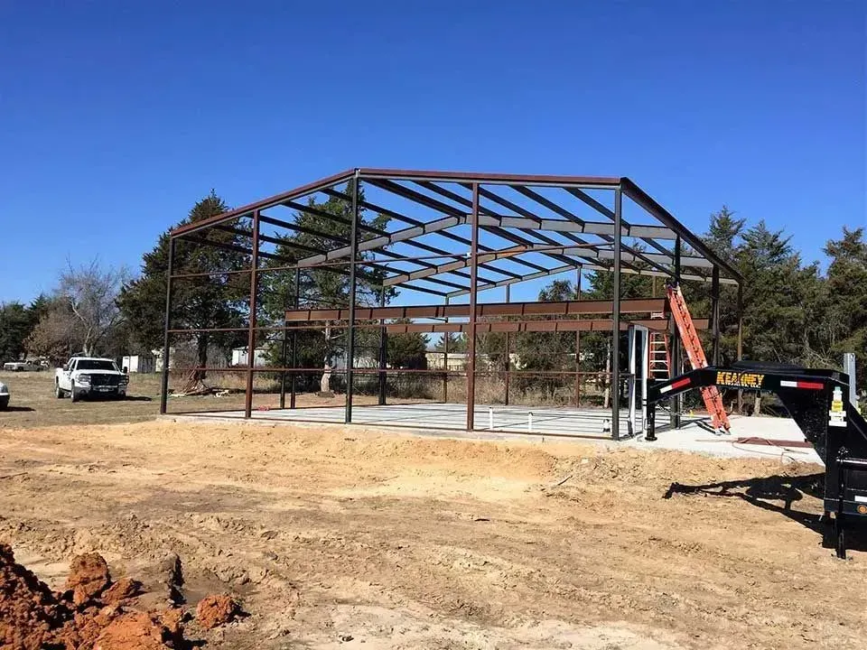 Steel frame of a building under construction on a concrete slab in an open, grassy area under a clear blue sky.