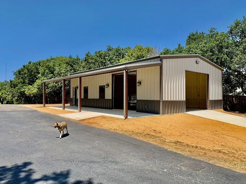 A tan and brown metal building with a covered porch and open garage, set beside a paved driveway with a dog walking by.