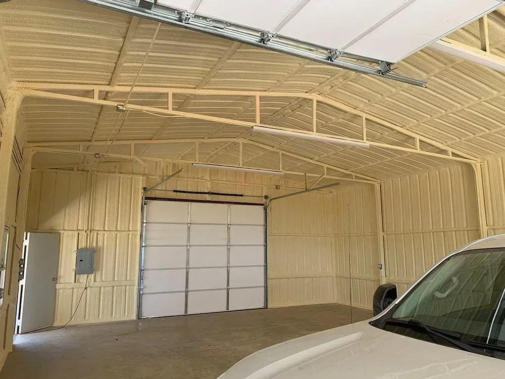 Interior of a large garage or workshop with beige foam-insulated walls and ceiling, a metal door, and a white vehicle.