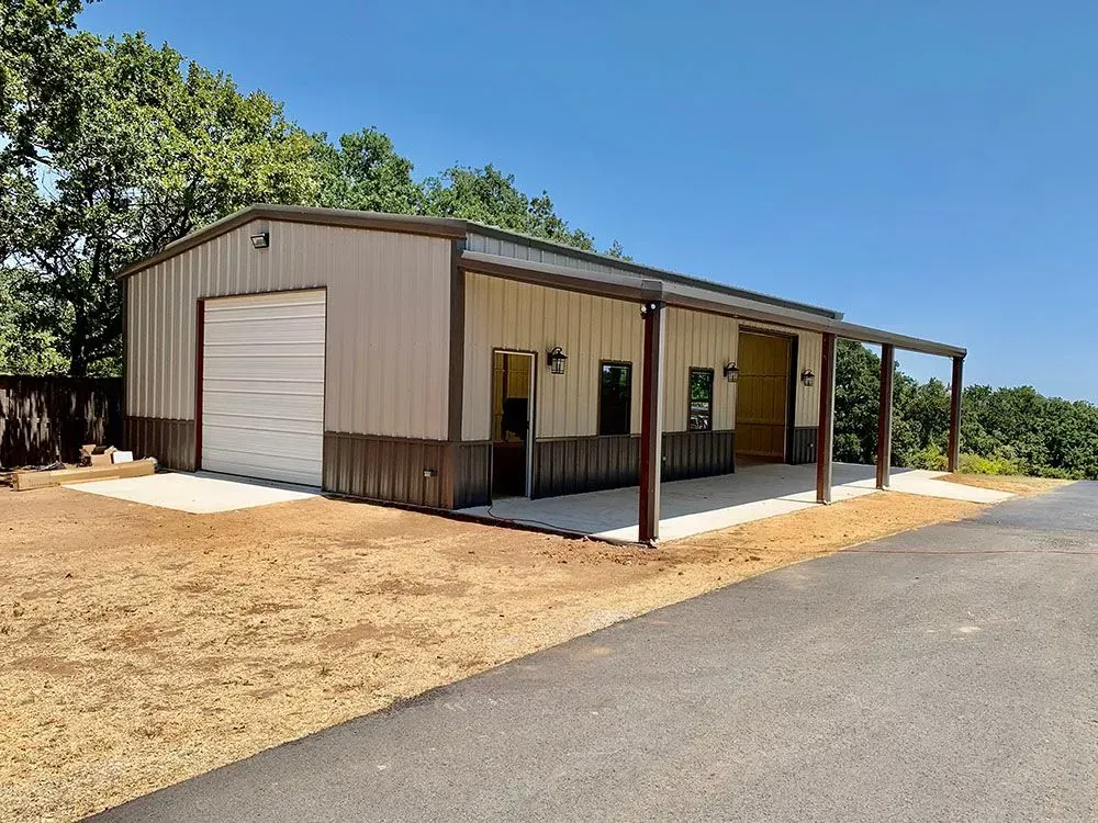 A tan metal barn with a roll-up door and covered side porch, situated next to a paved driveway and trees.