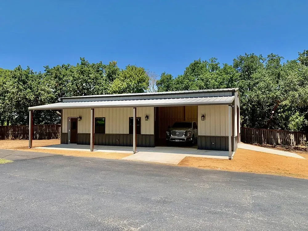 A modern metal building with a covered porch and open parking bay, set against a backdrop of trees under a clear sky.