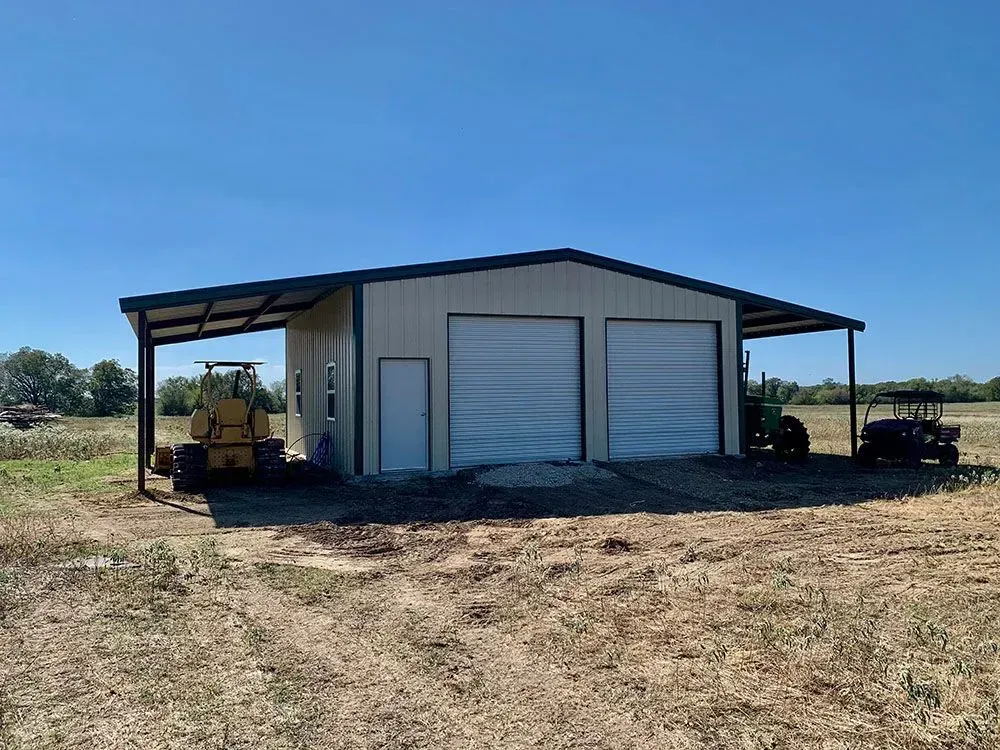 A tan metal building with two garage doors and a side door sits in a field with machinery parked under its side overhangs.