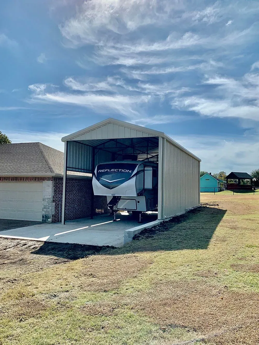 A large, off-white metal RV carport stands on a concrete pad in a grassy yard, housing a parked fifth-wheel trailer.